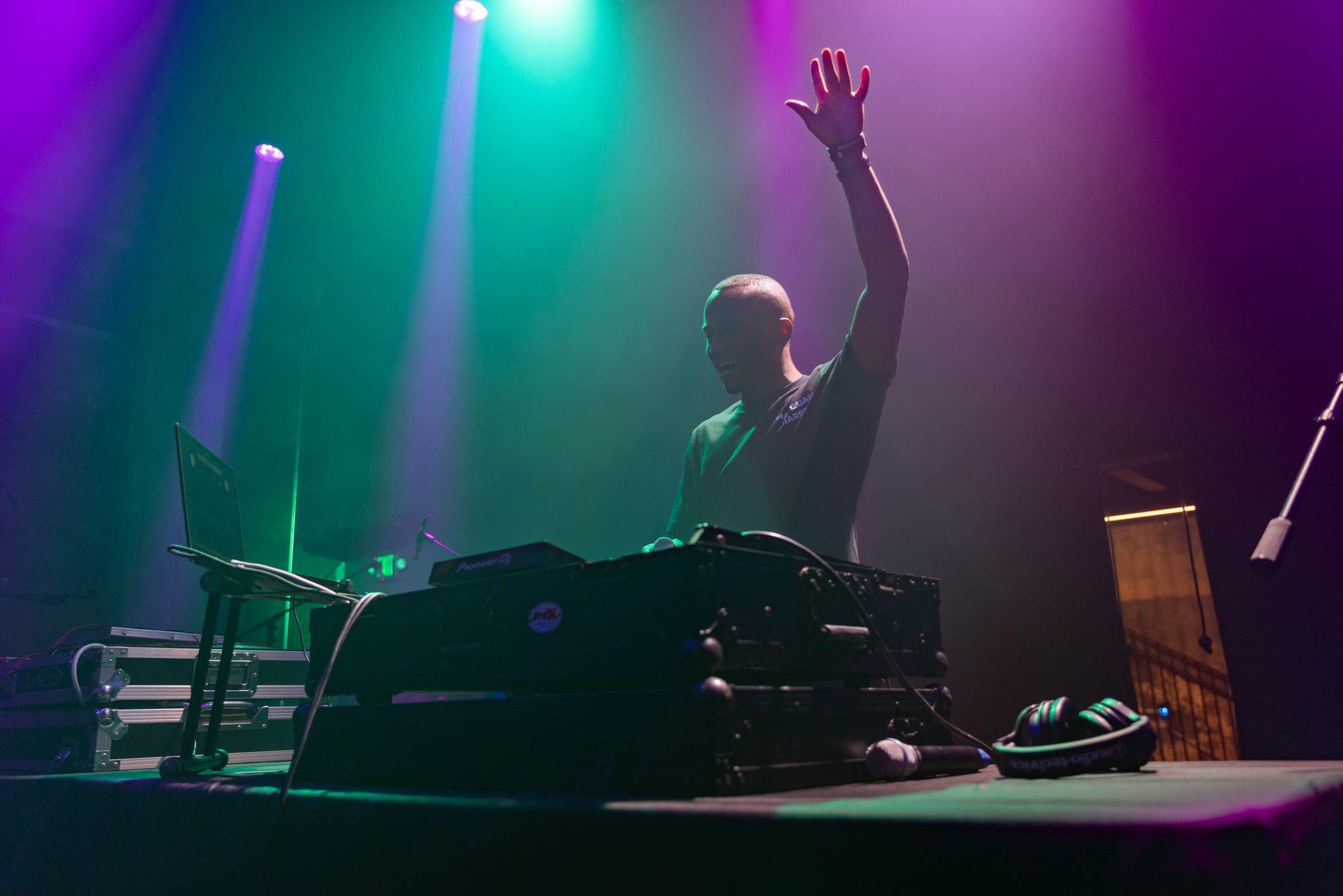 Wide view of MH at the DJ booth under teal and violet stage light, one arm raised