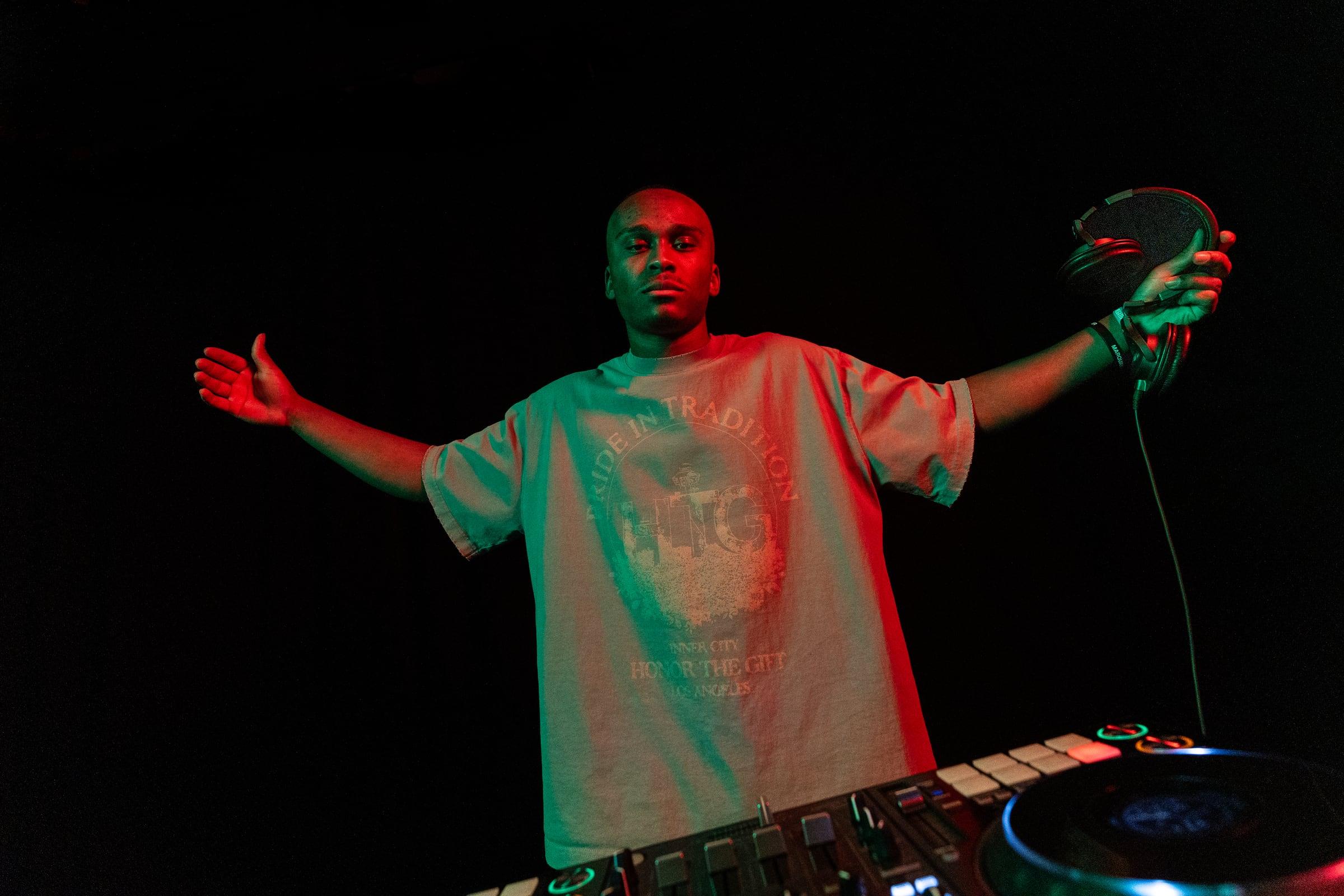 MH standing behind the decks with arms outstretched, headphones in one hand, under red stage light