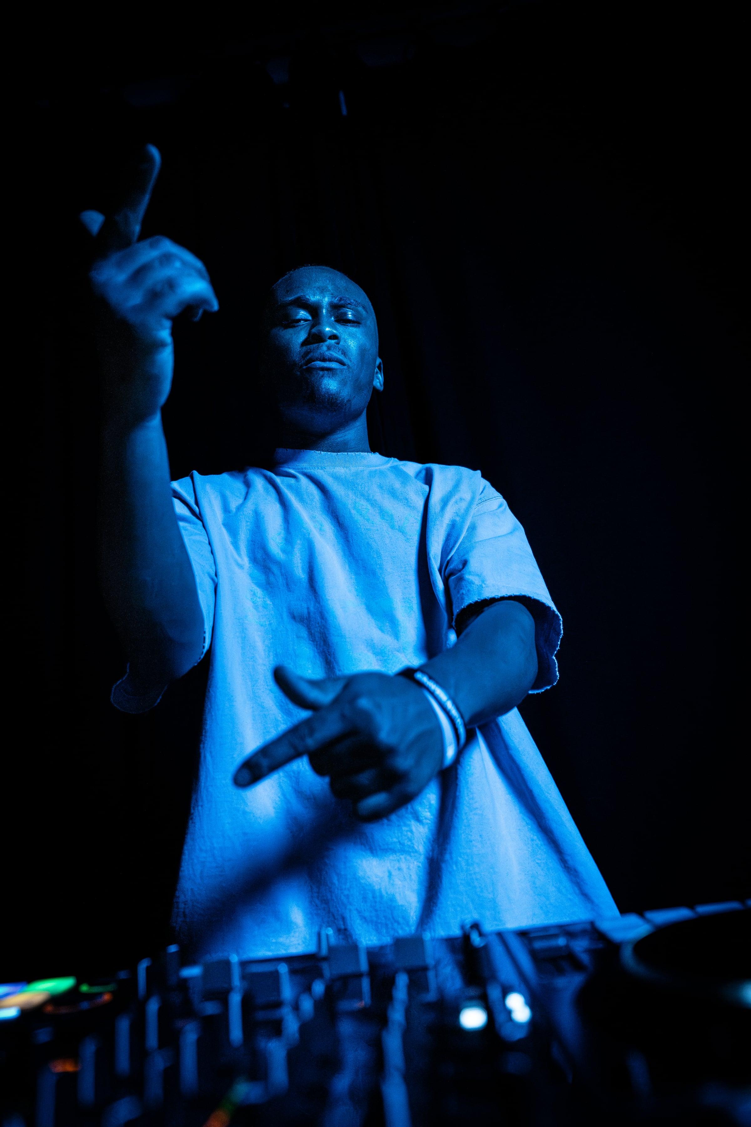 MH behind the decks under deep blue light, pointing mid-gesture