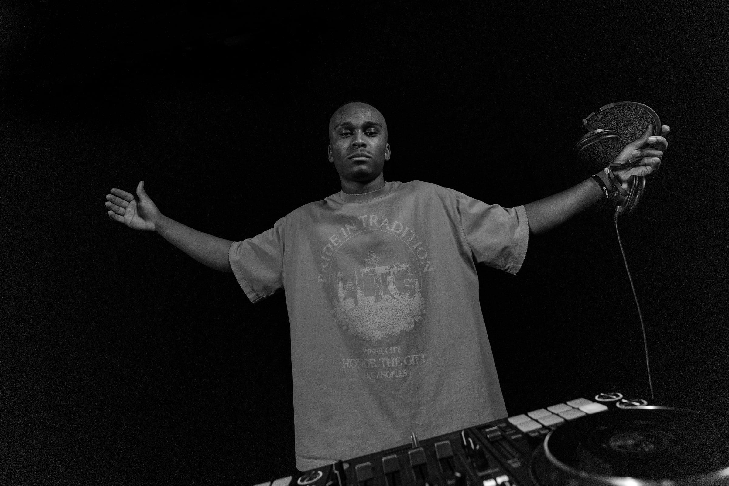 Black-and-white frame of MH behind the decks with arms outstretched
