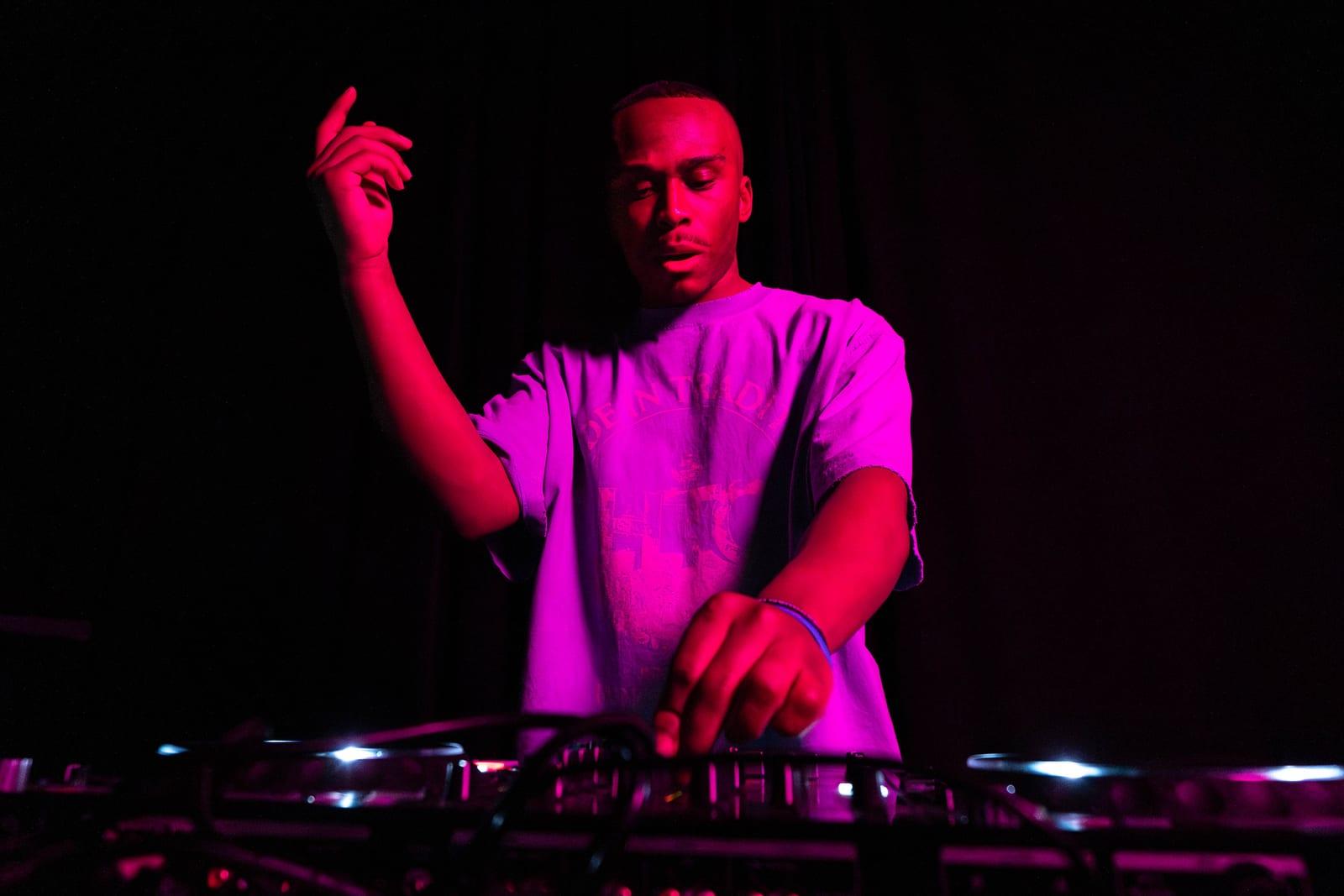 MH at the decks under magenta stage light, one arm raised