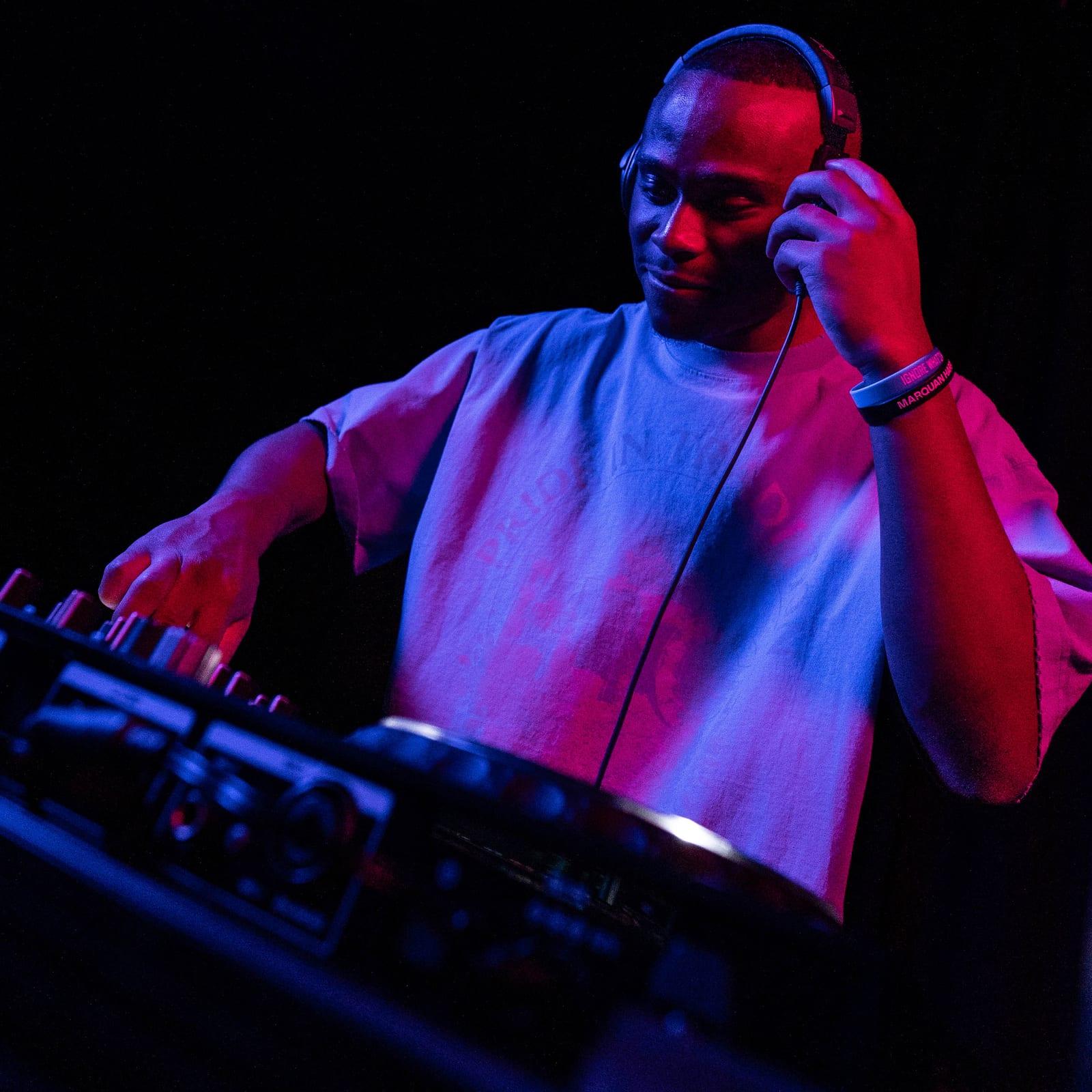 MH with one hand on his headphones and the other on the decks, lit in blue and magenta
