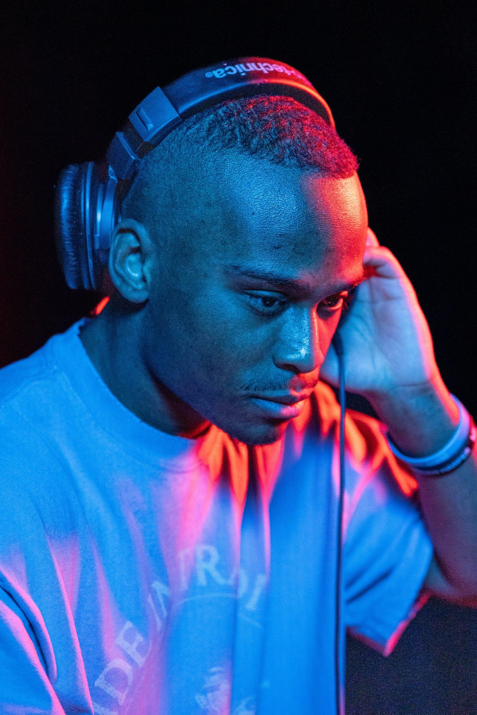 Portrait of MH with head down and one hand on his headphones, lit in blue and magenta